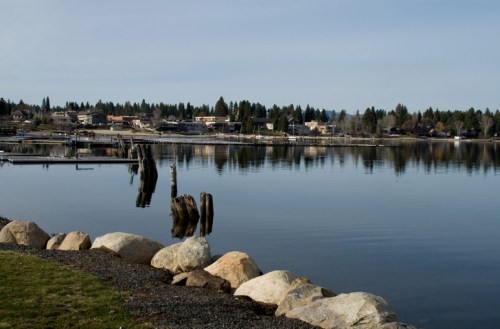 A calm lake reflecting a town with docks and trees, bordered by large rocks and grassy areas.
