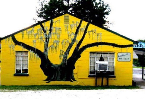 A bright yellow building with a large tree mural on the side, featuring whimsical figures hanging from the branches.