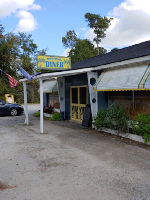 A diner with a blue exterior and yellow sign, featuring outdoor seating and surrounded by trees under a cloudy sky.