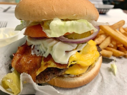 A close-up of a juicy burger with lettuce, tomato, pickles, onions, cheese, and bacon, served with fries on the side.