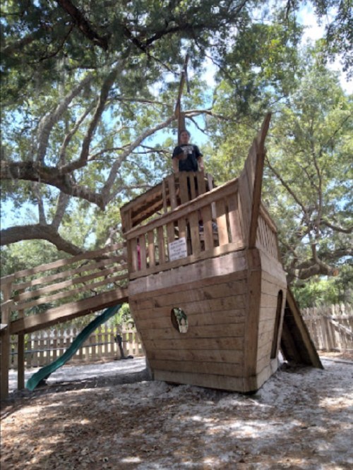 A wooden pirate ship play structure with a child standing on top, surrounded by trees and a slide.