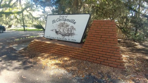 Sign for the Town of McClellanville, SC, displayed on a brick structure surrounded by trees.