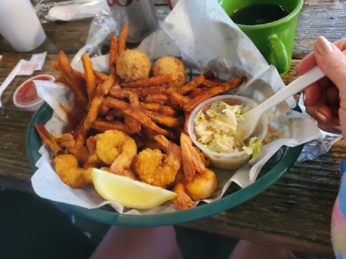 A plate of fried shrimp, sweet potato fries, hushpuppies, coleslaw, and a lemon wedge, with a drink in the background.
