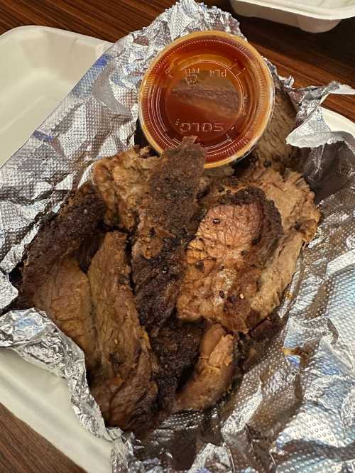 A container of sliced, smoky beef brisket wrapped in foil, accompanied by a small cup of barbecue sauce.