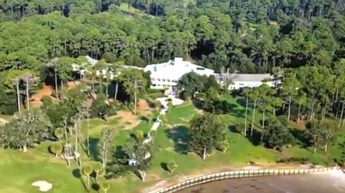 Aerial view of a large white mansion surrounded by lush greenery and a waterfront.