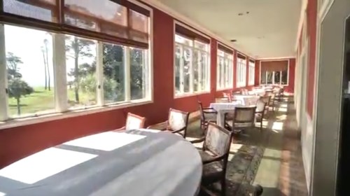 A sunlit dining area with round tables and chairs, featuring large windows overlooking a scenic view.