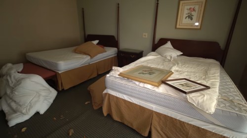 A disheveled hotel room with two unmade beds, a fallen blanket, and framed artwork on the floor.