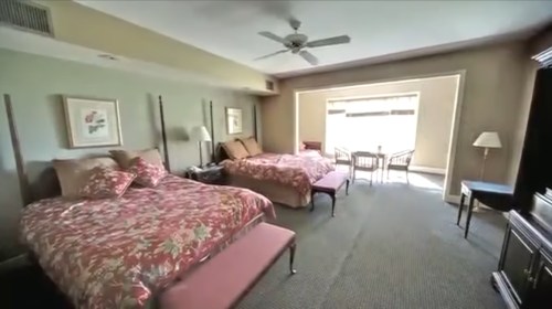A spacious hotel room with two beds, floral bedding, a seating area, and large windows letting in natural light.