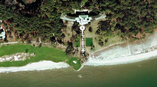 Aerial view of a large estate surrounded by trees, with a manicured lawn leading to a beach and water.
