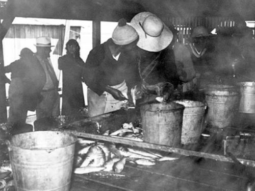 Black and white image of people preparing fish at a market, with buckets and smoke in a rustic setting.