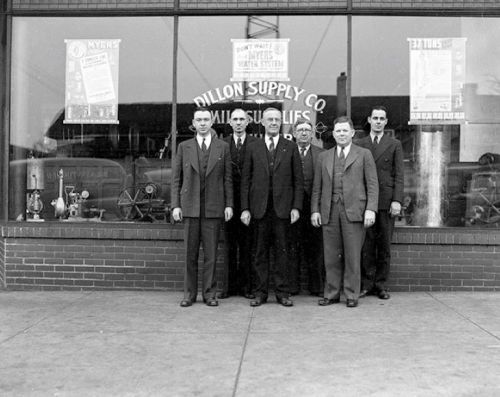 Black and white photo of six men in suits standing in front of a store window with "Dillon Supply Co." signage.
