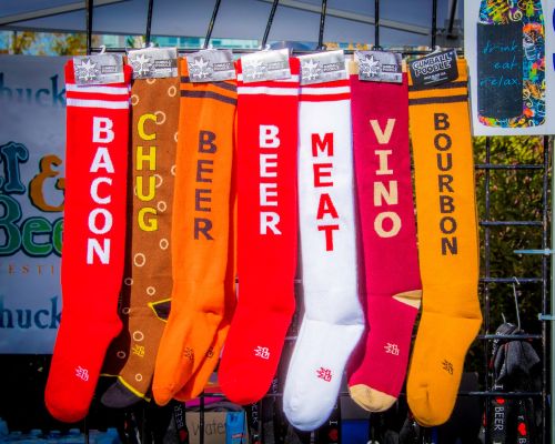 Colorful socks displayed at a festival, featuring fun food and drink-themed designs like "Bacon," "Beer," and "Bourbon."