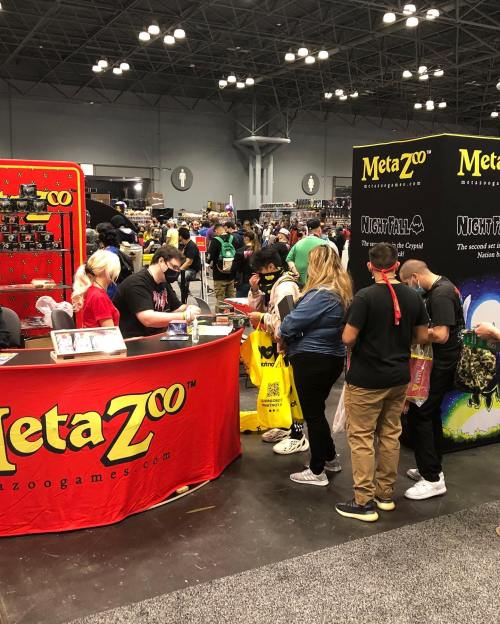 A busy convention scene with people gathered around a booth for MetaZoo Games, showcasing various products.