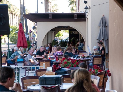 A bustling outdoor café scene with people dining and socializing under umbrellas and palm trees.