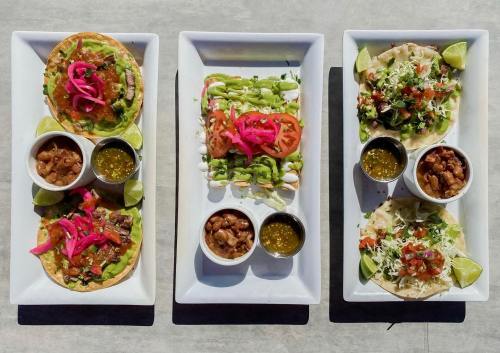 Three plates of tacos with various toppings, garnished with lime, pickled onions, and sauces on a gray surface.