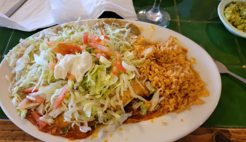 A plate of enchiladas topped with lettuce, tomatoes, and sour cream, served with rice and a side of guacamole.