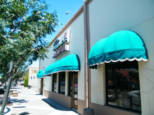 Exterior of a building with teal awnings and large windows, set along a sidewalk lined with trees.