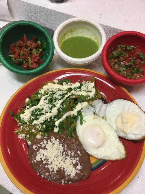 A colorful plate of enchiladas topped with cheese, eggs, and fresh herbs, accompanied by salsa and green sauce in bowls.
