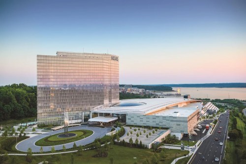A modern hotel and casino complex with a glass facade, surrounded by landscaped gardens and a river in the background.