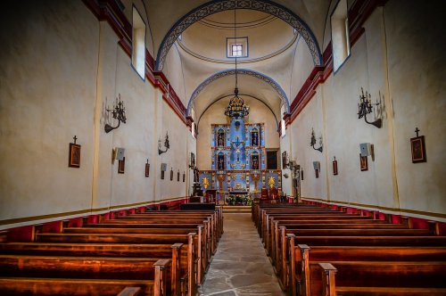 Interior of a church featuring wooden pews, a blue altar, and decorative chandeliers under a high arched ceiling.