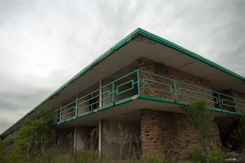 Abandoned building with a green roof and balcony, surrounded by overgrown vegetation under a cloudy sky.