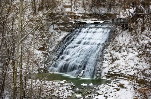 A snowy waterfall cascades down rocky cliffs, surrounded by trees and a serene winter landscape.