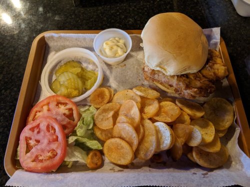 A tray with a fried chicken sandwich, pickles, sliced tomatoes, lettuce, and potato chips with a side of mayonnaise.