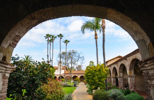 A view through an archway showcasing a garden with palm trees and historic buildings under a blue sky.