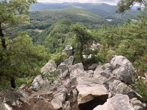 A rocky outcrop overlooks a lush green valley and distant mountains under a cloudy sky.