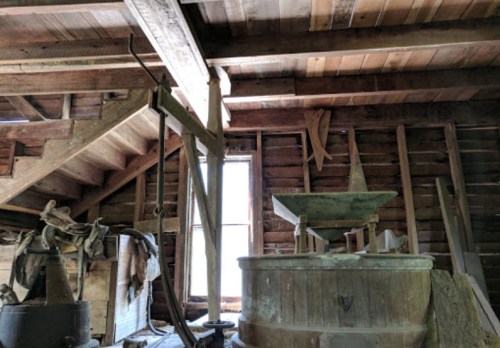 Interior of an old barn with wooden beams, a staircase, and vintage farming equipment near a window.