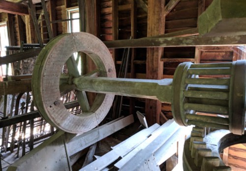 Close-up of a large wooden wheel and gears inside a rustic, dimly lit barn.