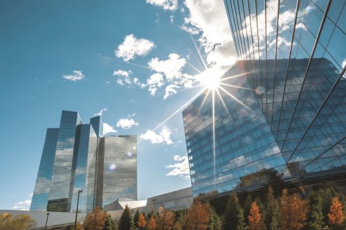 Sunlight reflects off modern glass skyscrapers against a blue sky with scattered clouds and autumn trees in the foreground.