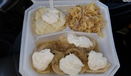 A takeout container with mashed potatoes, noodles, and dumplings topped with sour cream and onions.