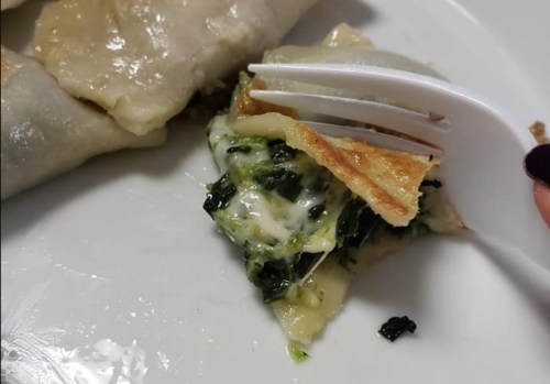 A close-up of a fork lifting a piece of stuffed dumpling filled with greens and cheese on a white plate.