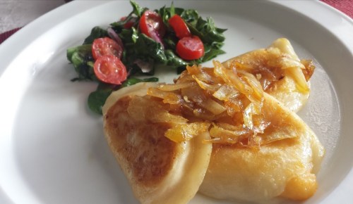 Plate of pierogi topped with caramelized onions, served with a side salad of spinach and cherry tomatoes.