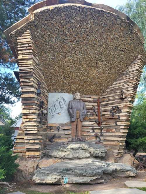 A wooden structure with stacked logs features a statue of a man standing on a rock, surrounded by tools and artwork.