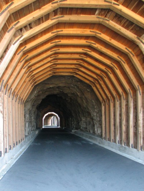 A wooden tunnel with a curved ceiling, leading into a rocky passageway, illuminated by light at the far end.