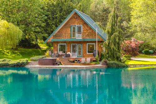 A charming wooden cabin by a serene turquoise lake, surrounded by lush greenery and ducks swimming nearby.
