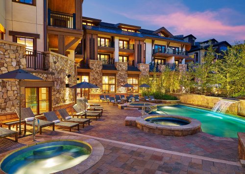 A luxurious outdoor pool area with hot tubs, lounge chairs, and stone buildings at sunset.