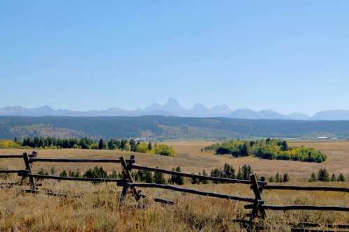 A scenic view of mountains in the distance, framed by a wooden fence and grassy fields under a clear blue sky.