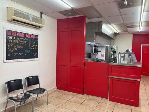 Interior of a casual eatery featuring a red wall, menu board, kitchen area, and two empty chairs.
