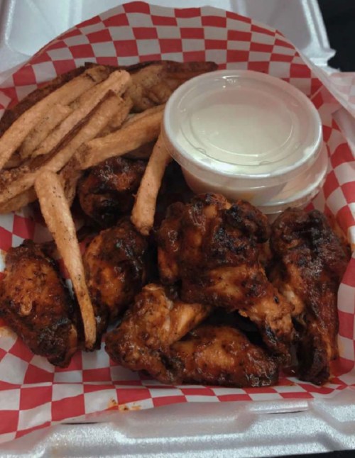 A takeout container with crispy chicken wings, seasoned fries, and a small cup of dipping sauce on a red checkered paper.