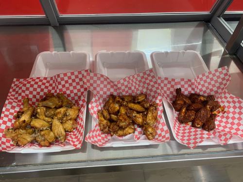 Three trays of chicken wings in red and white checkered paper, each with a different sauce, displayed in a glass case.