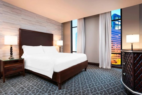 A modern hotel room featuring a king bed, bedside tables, and large windows with a city view at dusk.