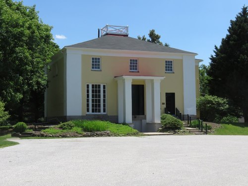 A two-story house with a yellow and white facade, featuring large windows and a front porch, surrounded by greenery.