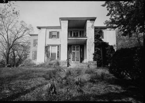 A large, historic house with a central entrance, surrounded by overgrown grass and trees.