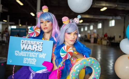 Two colorful performers with pastel hair and candy-themed outfits hold a sign that reads "Dessert Wars: Sweetest Day Ever."