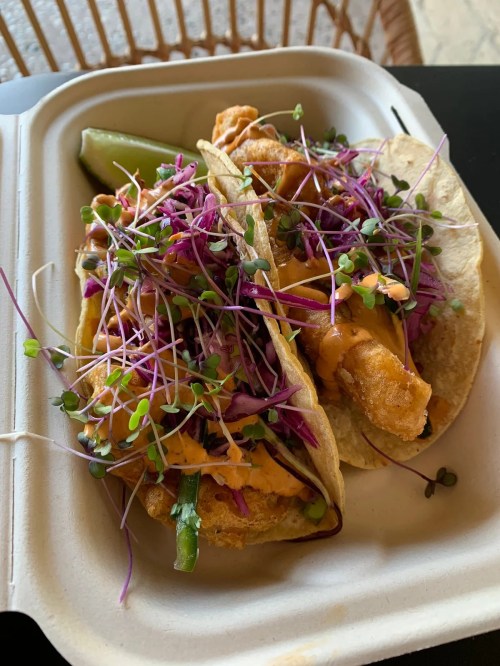 Two tacos filled with crispy fish, topped with purple cabbage, microgreens, and a drizzle of sauce, served with a lime wedge.