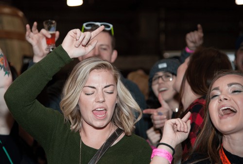 A lively crowd enjoys music, with a woman in the foreground singing and dancing, surrounded by enthusiastic friends.