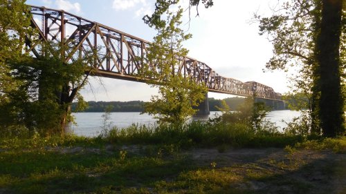 A rusted metal bridge spans a river, framed by trees and bathed in warm sunlight.